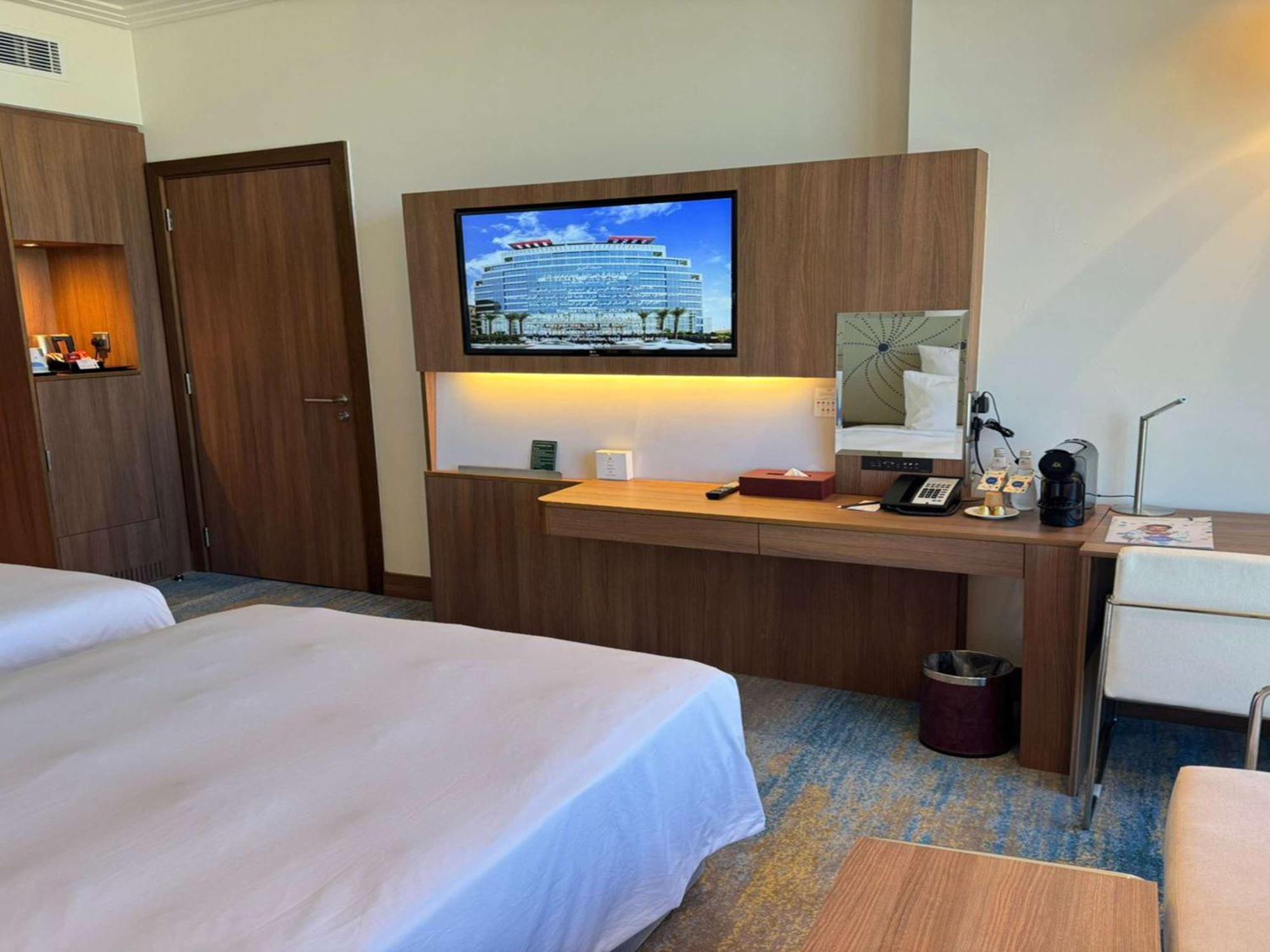 Novotel Jazan Extérieur photo The photo shows a hotel room with two beds made up with white linens. There is a wooden wall unit that features a flat-screen television mounted on it, displaying a picture of a large building. Below the TV, there is a desk area with various items, i